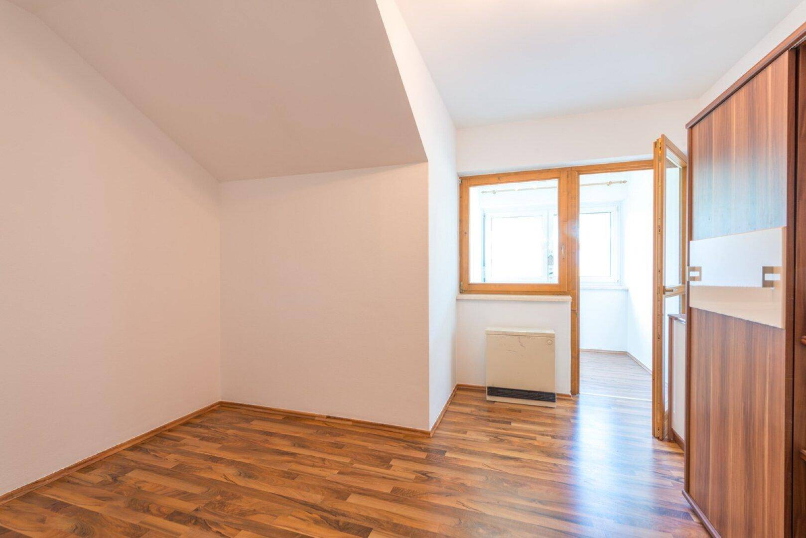 Helles Zimmer mit Dachschräge, Laminatboden und einem Fenster mit Blick ins Grüne.