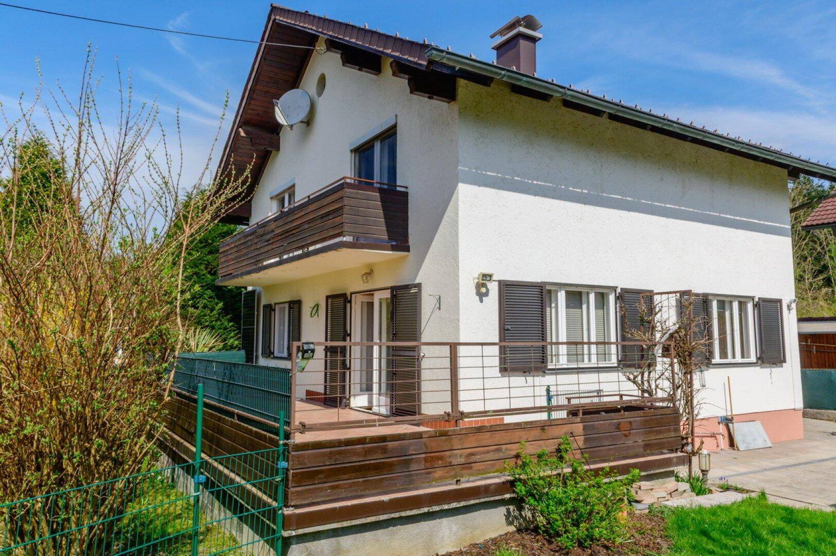 Charmantes Einfamilienhaus mit weißer Fassade, Holzterrasse und Balkon, umgeben von viel Grün.