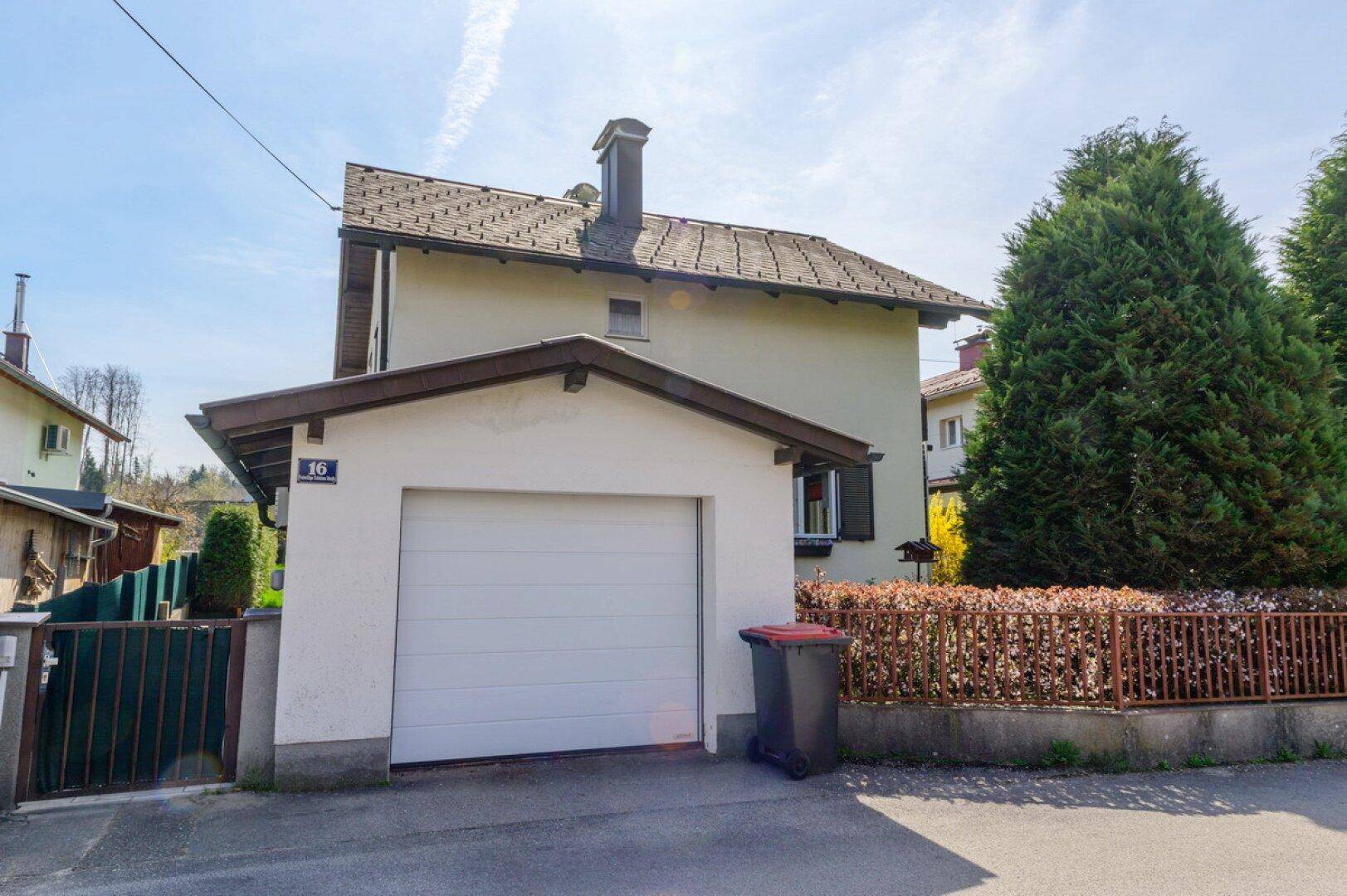 Rückansicht eines Einfamilienhauses mit integrierter Garage und Zufahrt, umgeben von Hecken und Bäumen.