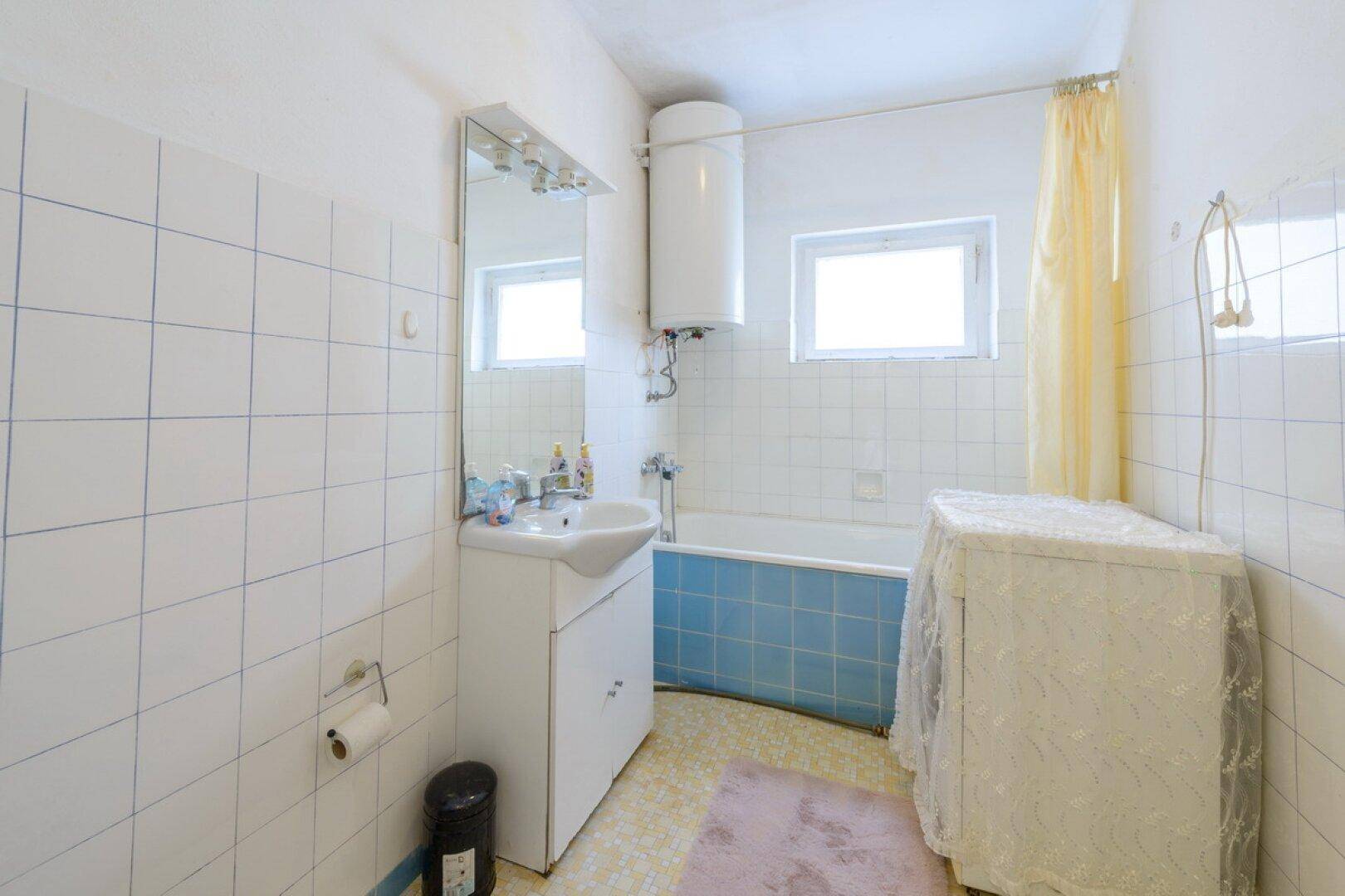Badezimmer im Stil der 80er Jahre mit Badewanne, Waschbecken und weißen Fliesen an den Wänden.