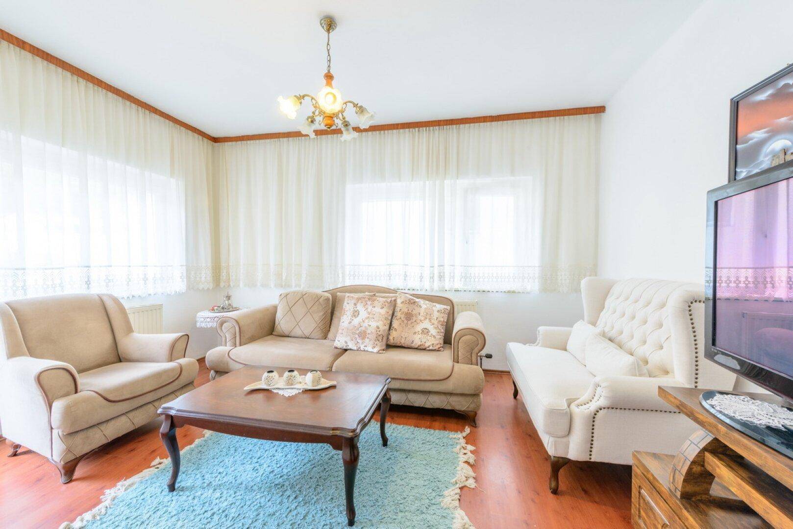 Helles Wohnzimmer mit beigen Sofas, Holzboden und großen Fenstern, die viel Tageslicht hereinlassen.