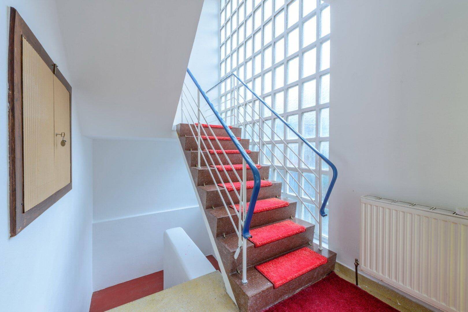 Treppenhaus mit Glasbausteinwand, die für natürliches Licht sorgt, und roten Teppichstufen.