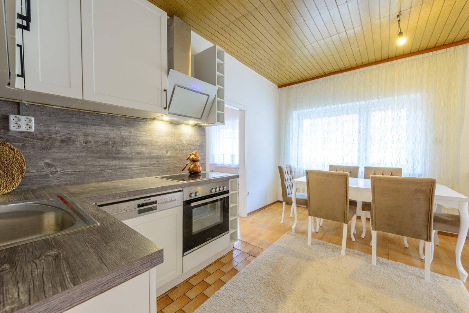 Moderne Einbauküche mit weißen Schränken, integrierten Geräten und einem Essbereich mit Holztisch.