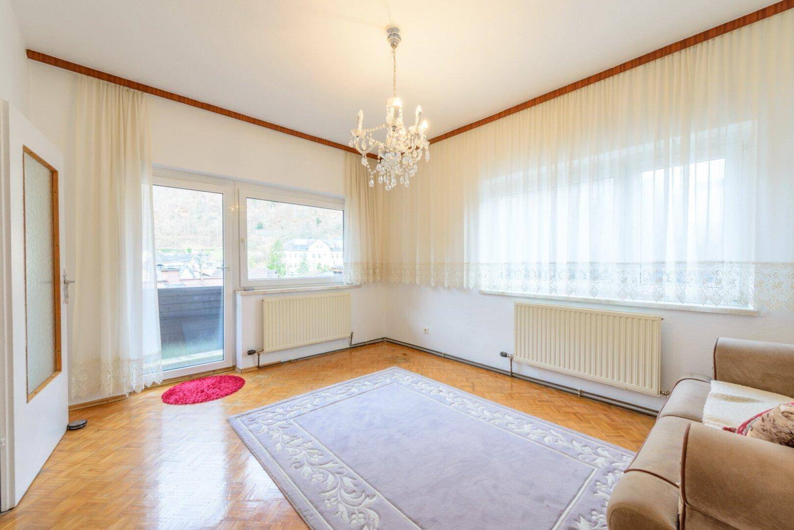 Heller Raum mit Holzboden, Zugang zu einem Balkon und Blick auf die umliegende Berglandschaft.
