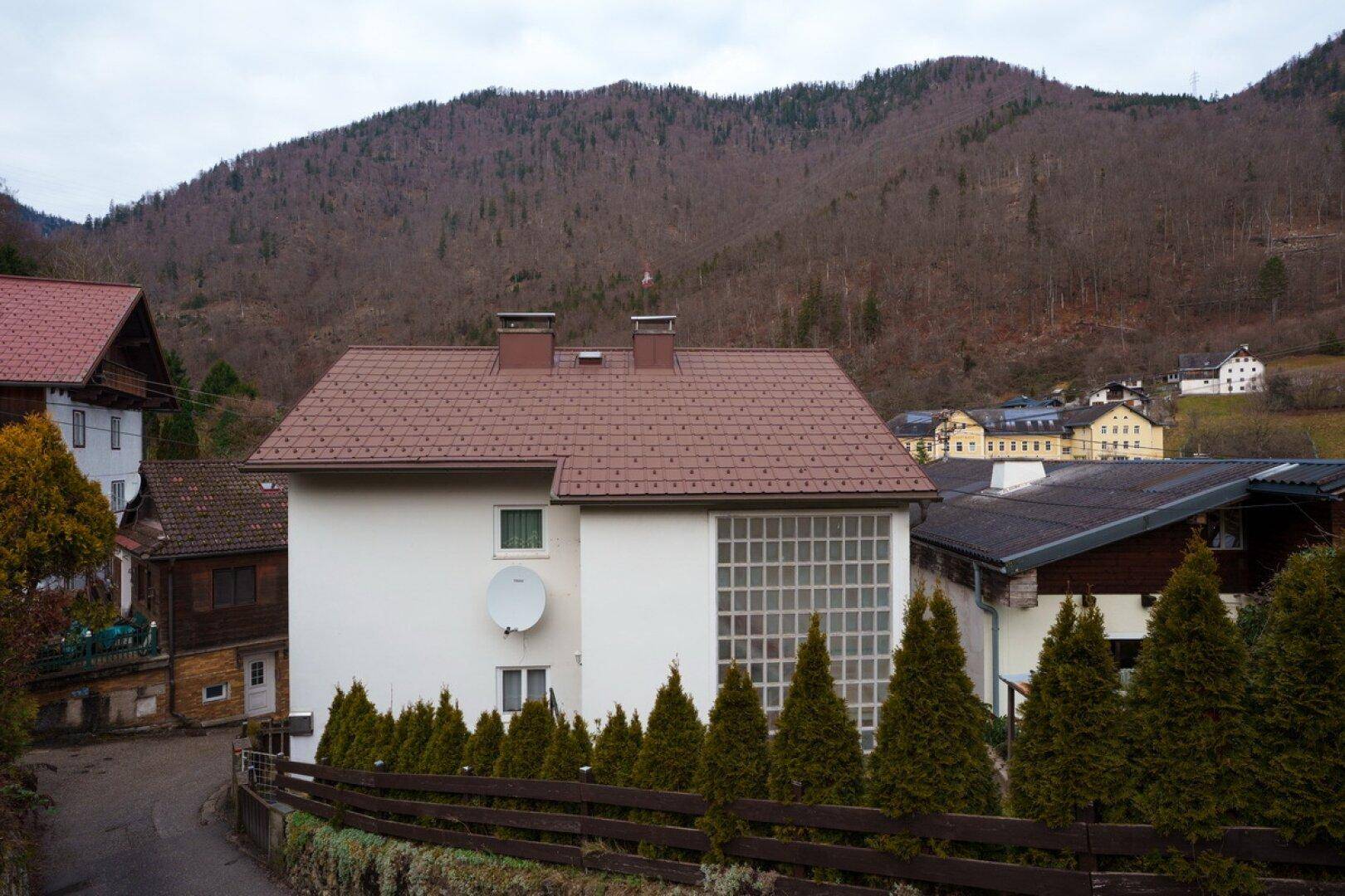 Ansicht des Wohnhauses mit braunem Satteldach und einem Satellitenempfänger vor einer bewaldeten Berglandschaft.