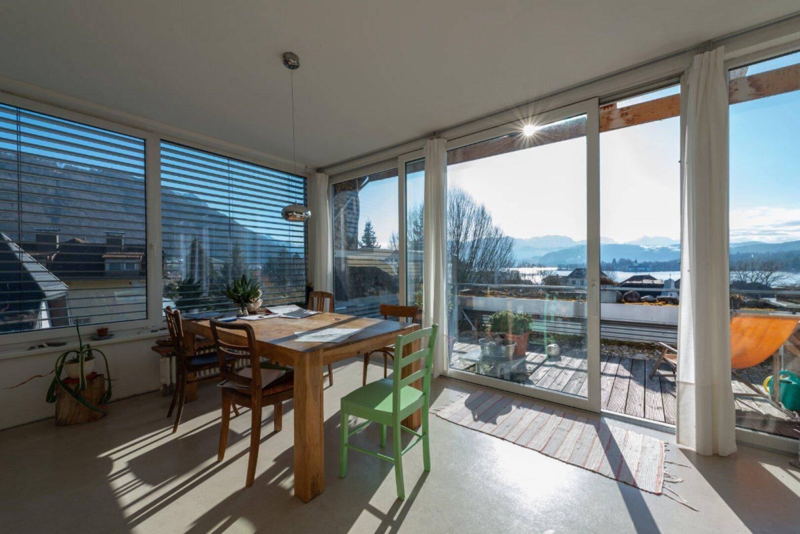 Sonniger Essbereich mit Holztisch, großen Fenstern und weitem Blick auf den See und die Berge.