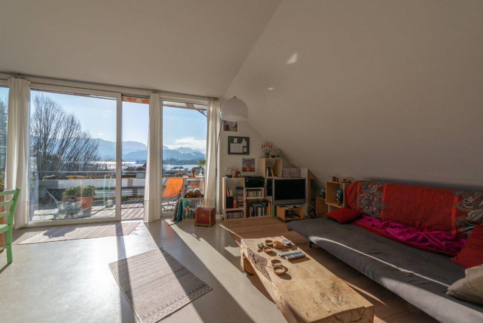 Helles Wohnzimmer im Dachgeschoss mit großen Fenstern, Seeblick und Zugang zum Balkon.