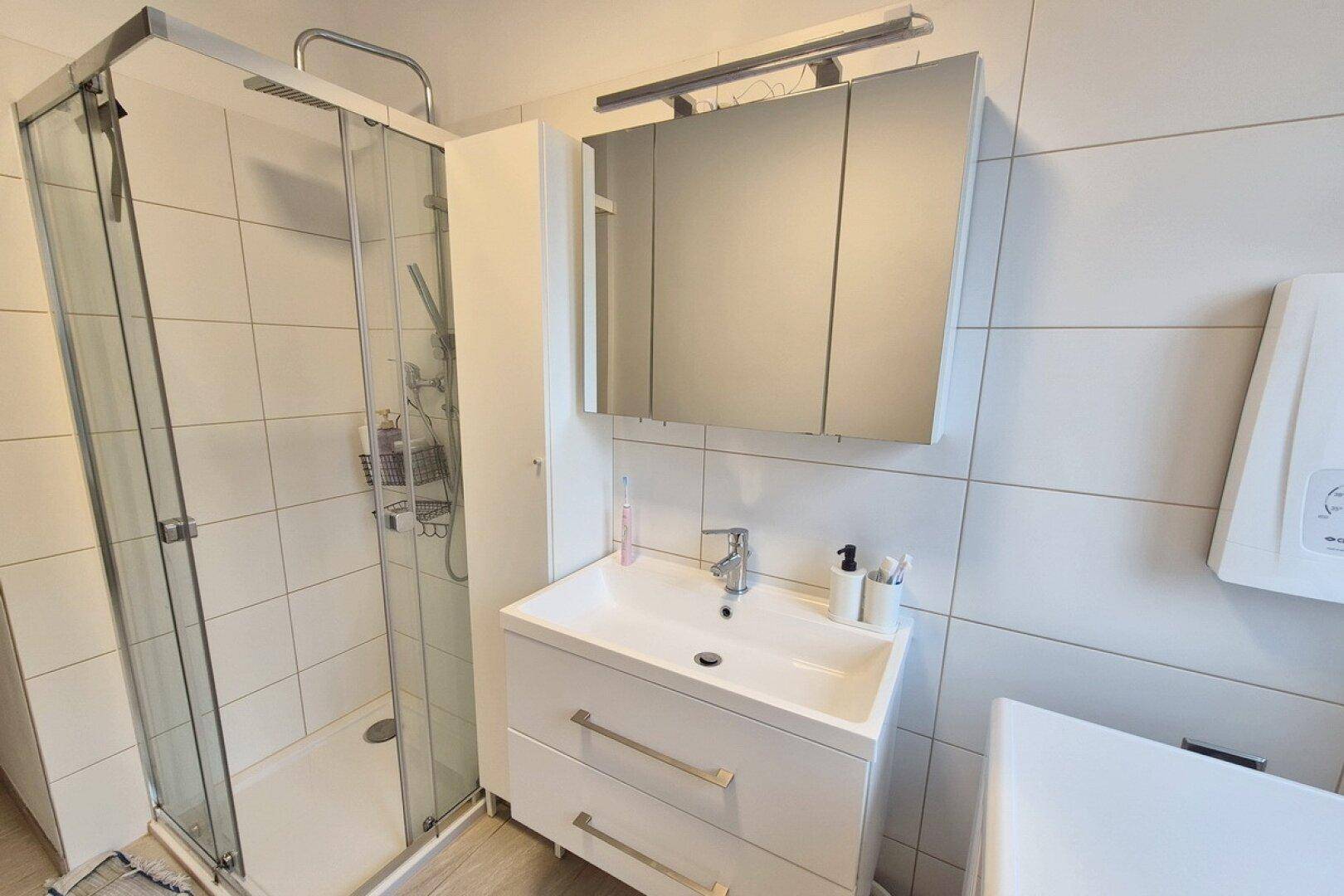 Modernes Badezimmer mit Duschkabine, Waschbecken und Spiegelschrank, hell gefliest.