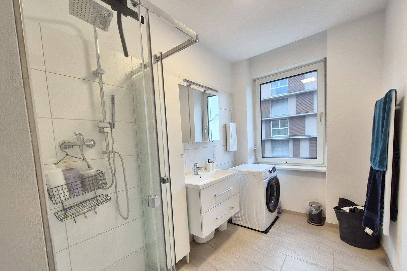 Helles Badezimmer mit Duschkabine, Waschbecken und Waschmaschine, gut beleuchtet.