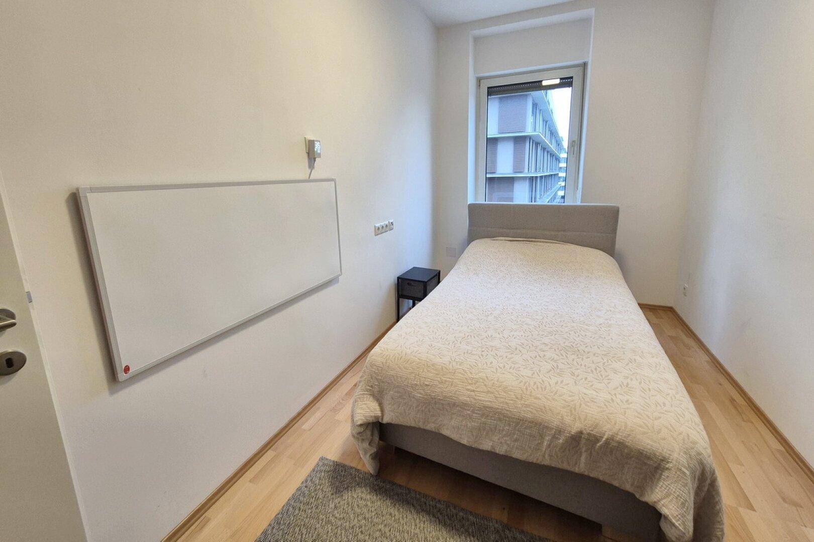 Helles Schlafzimmer mit Einzelbett, Holzfußboden und einem Fenster mit Blick nach draußen.