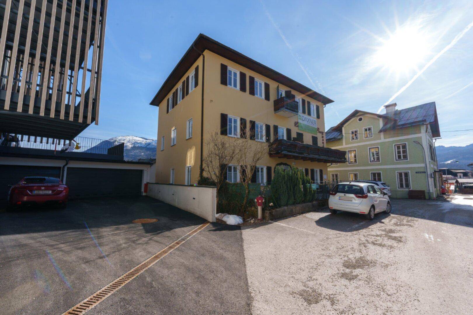 Gebäudeansicht der ehemaligen Pension mit Parkplätzen und umliegender Berglandschaft.