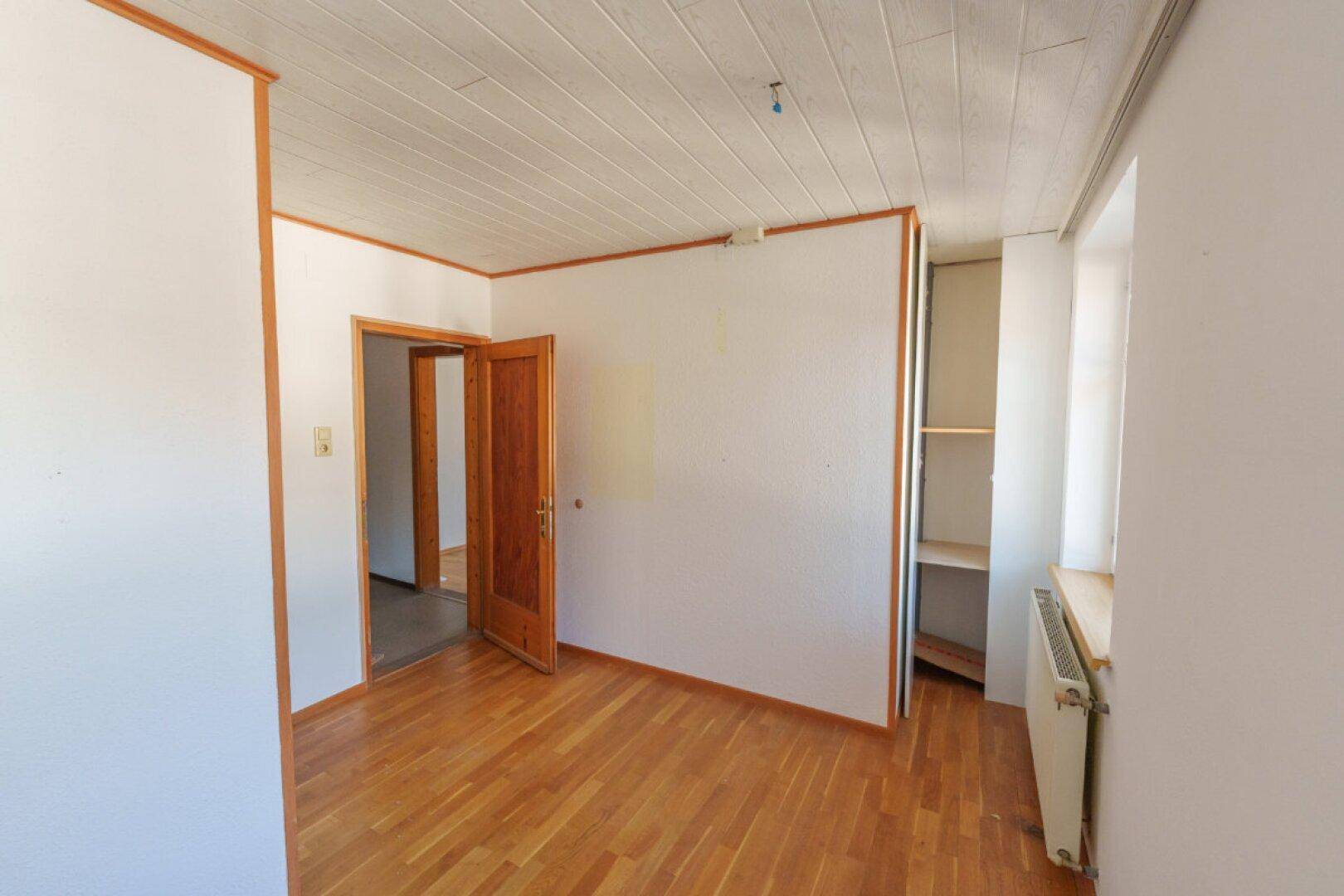 Einfaches Zimmer mit Holzboden, weiß gestrichenen Wänden und offenem Schrankbereich.
