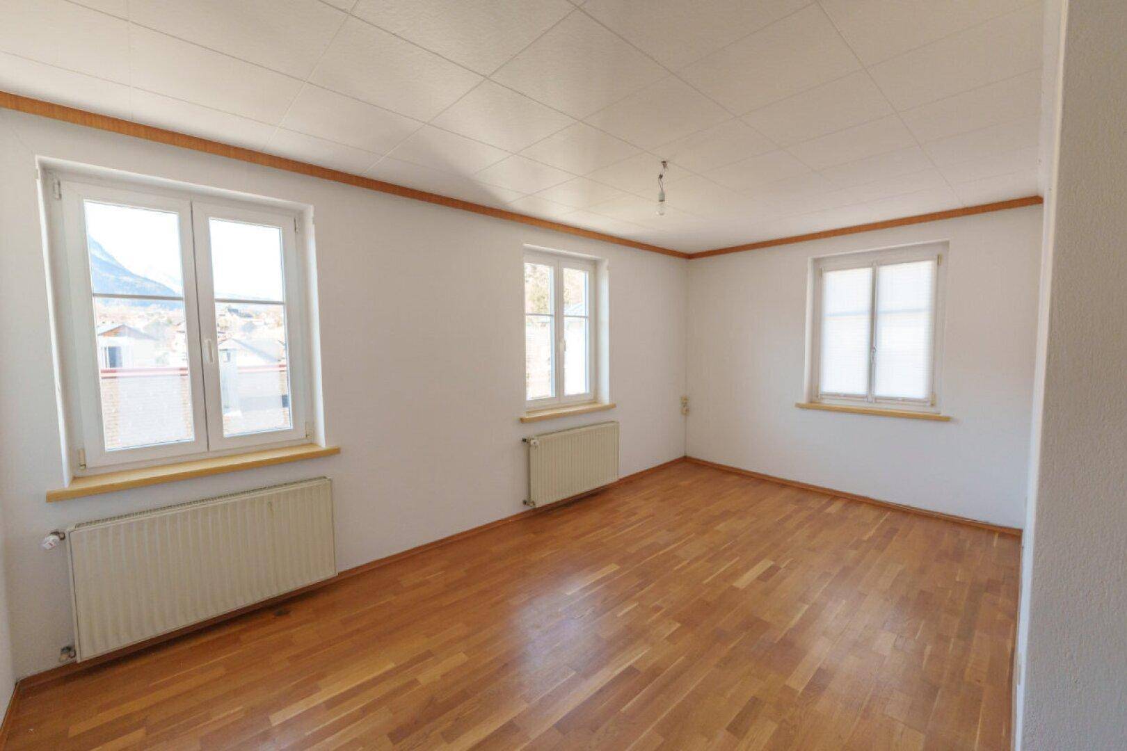 Helles Zimmer mit Holzboden, weißen Wänden und mehreren Fenstern.