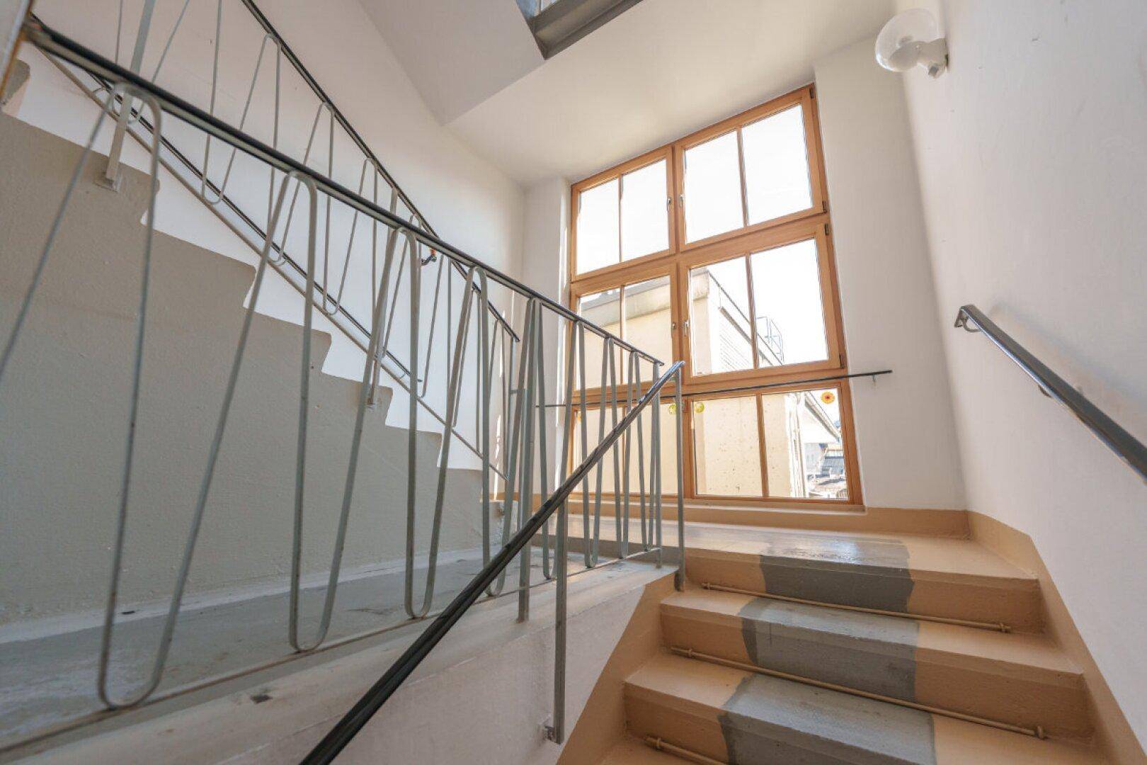 Treppenhaus mit Holzfenstern und Metallgeländer, das viel Tageslicht hereinlässt.
