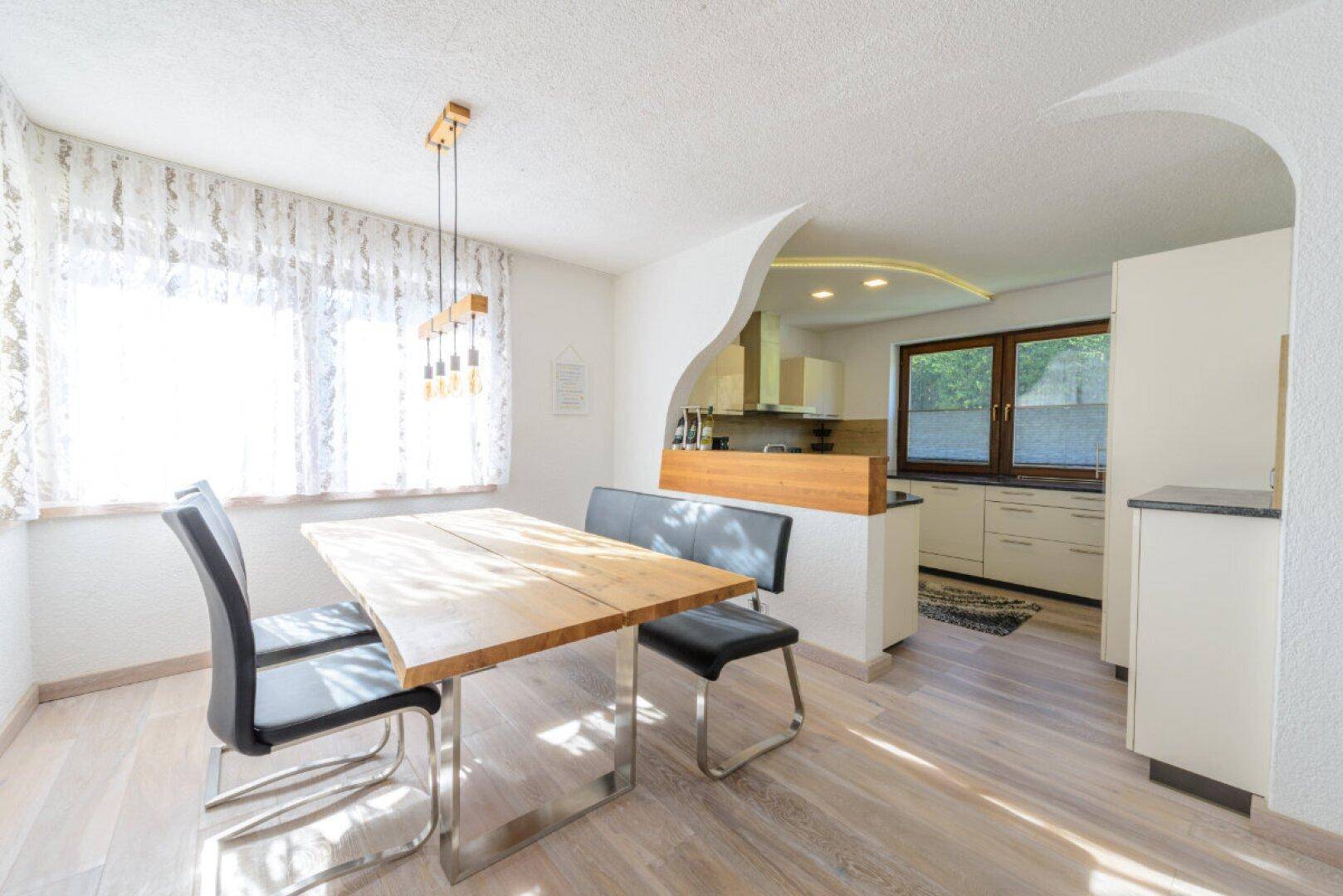 Helle Küche mit Essbereich, Holztisch und Blick ins Grüne durch große Fenster.