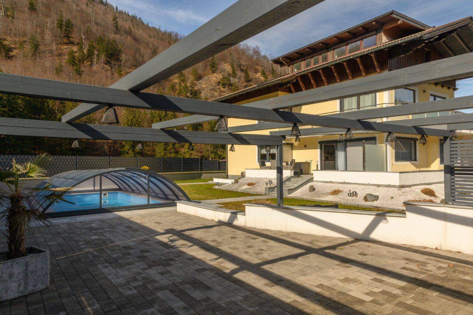 Außenbereich mit überdachtem Pool, Pergola und Blick auf die bewaldeten Berge.