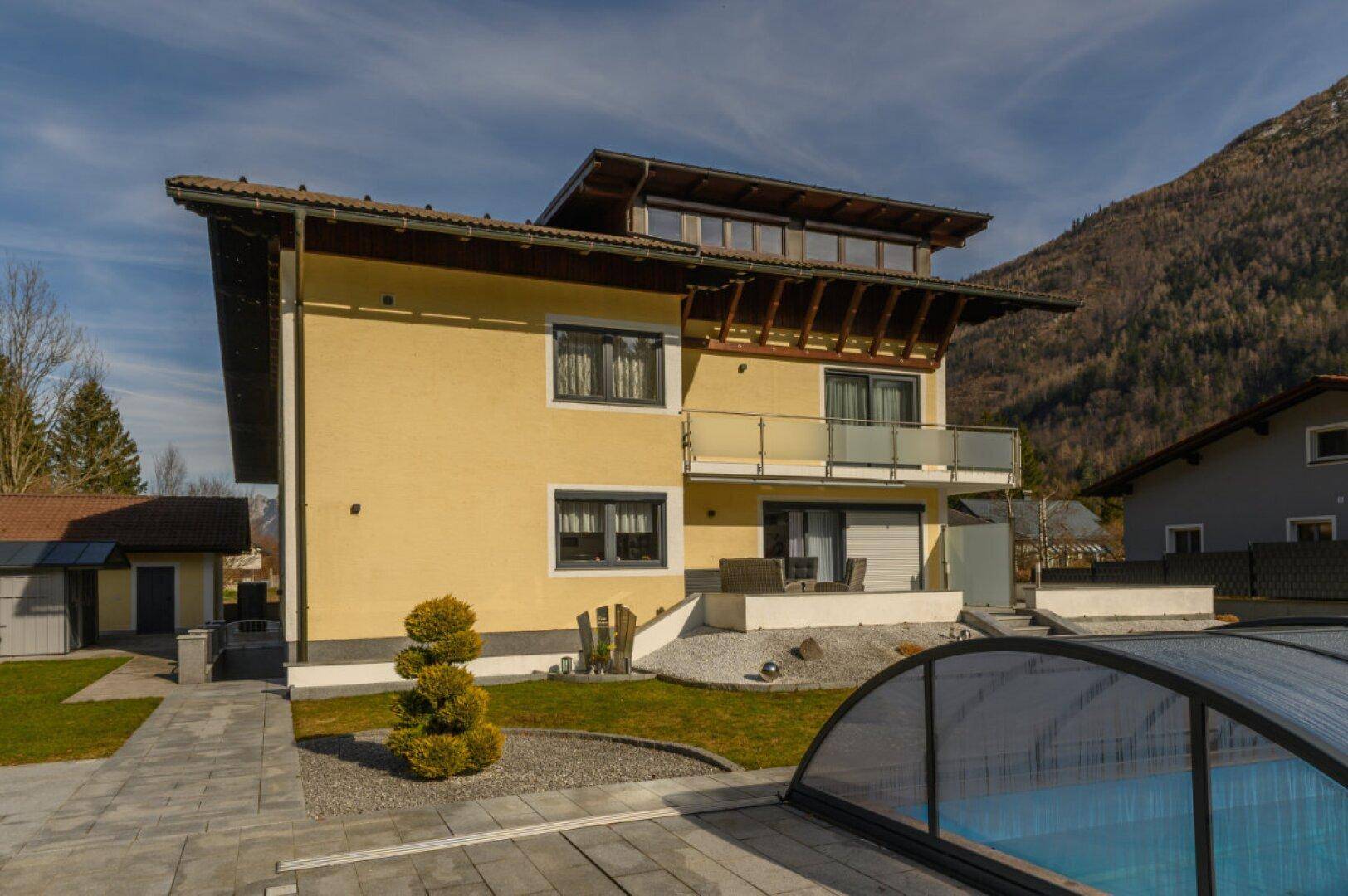Rückansicht des Hauses mit Garten, überdachtem Pool und Blick auf die bewaldeten Berge.
