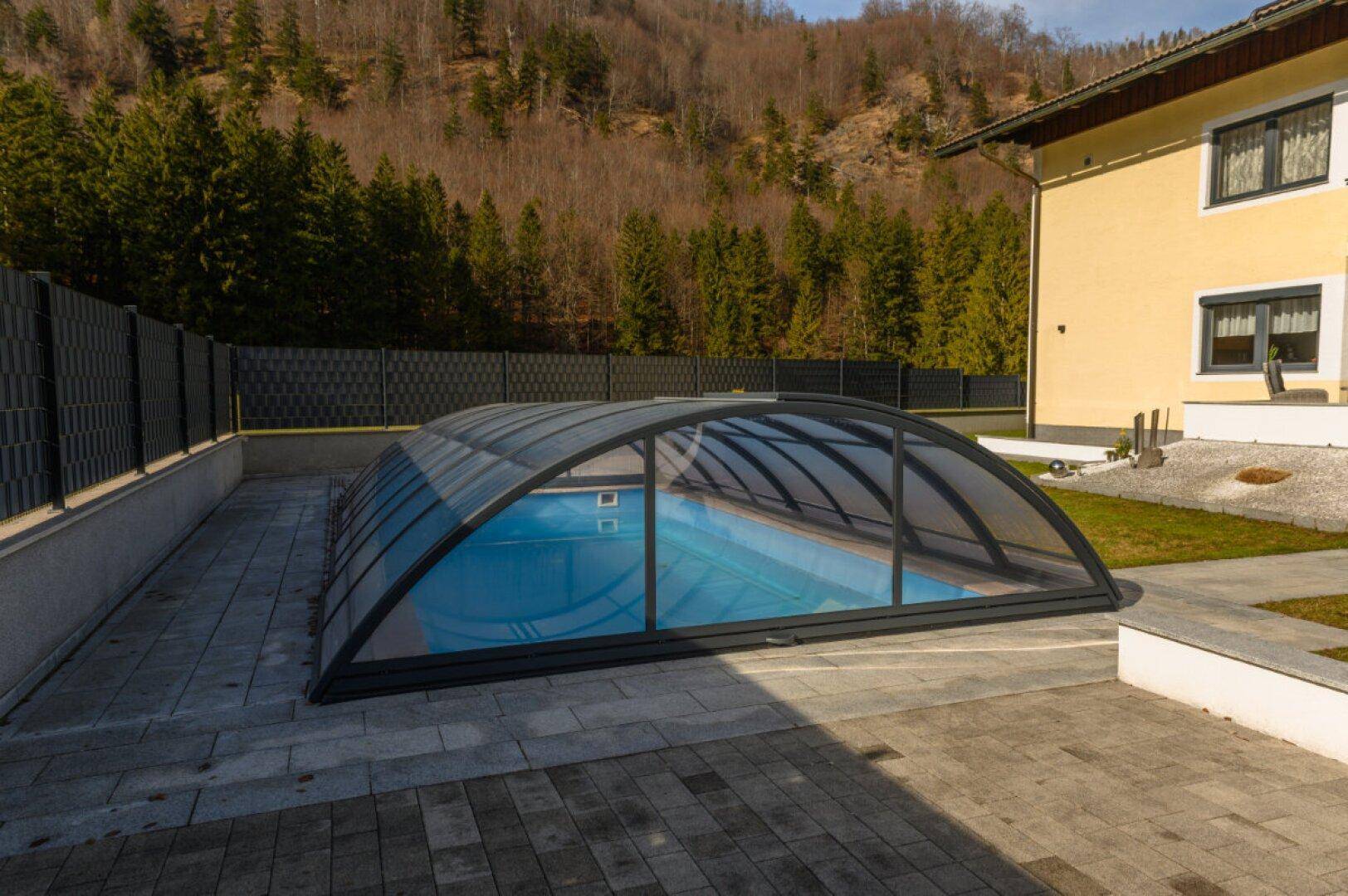 Überdachter Swimmingpool im Gartenbereich mit gepflasterter Fläche und Zaun.