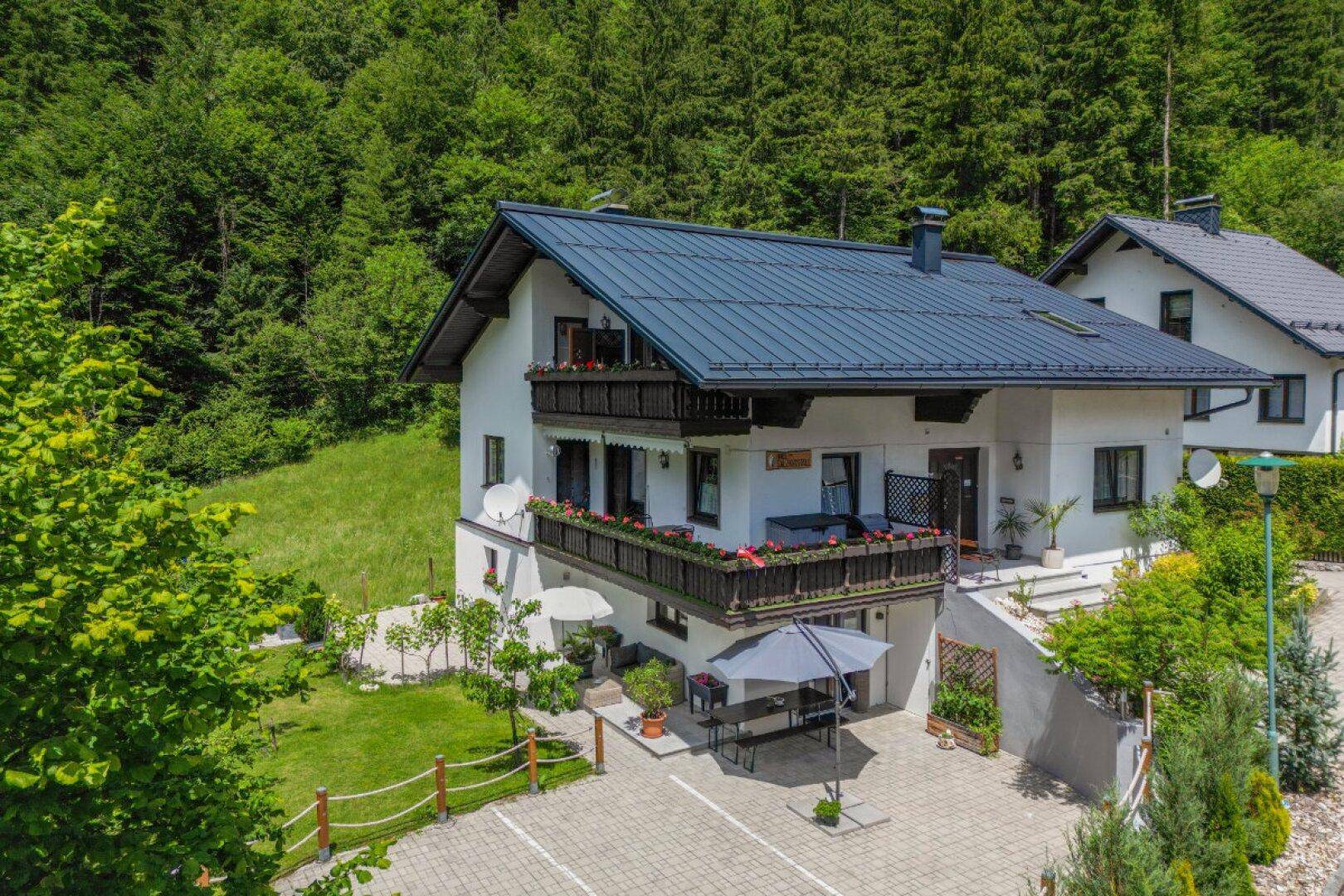 Luftaufnahme eines Hauses mit dunklem Dach, Balkonen und gepflastertem Vorplatz vor bewaldeten Bergen.