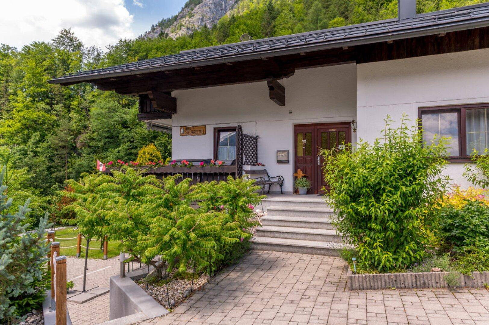 Eingangsbereich des Hauses mit gepflastertem Weg, Stufen und gepflegten Büschen vor grüner Kulisse.