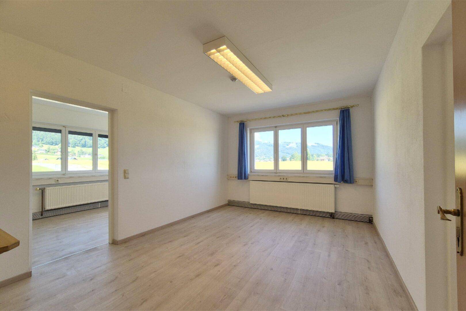Helles Bürozimmer mit Laminatboden, blauen Vorhängen und einem weiten Blick auf die umliegende Landschaft.