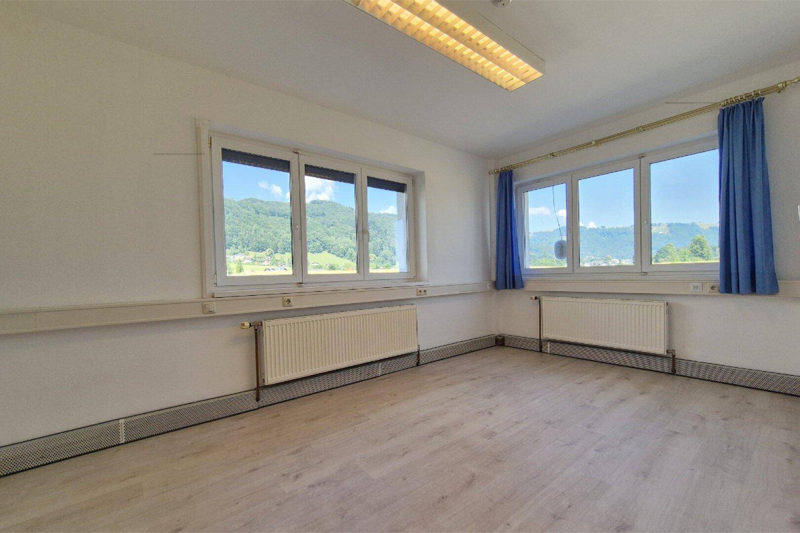 Helles und leeres Bürozimmer mit Laminatboden und zwei großen Fenstern, die viel Tageslicht hereinlassen.