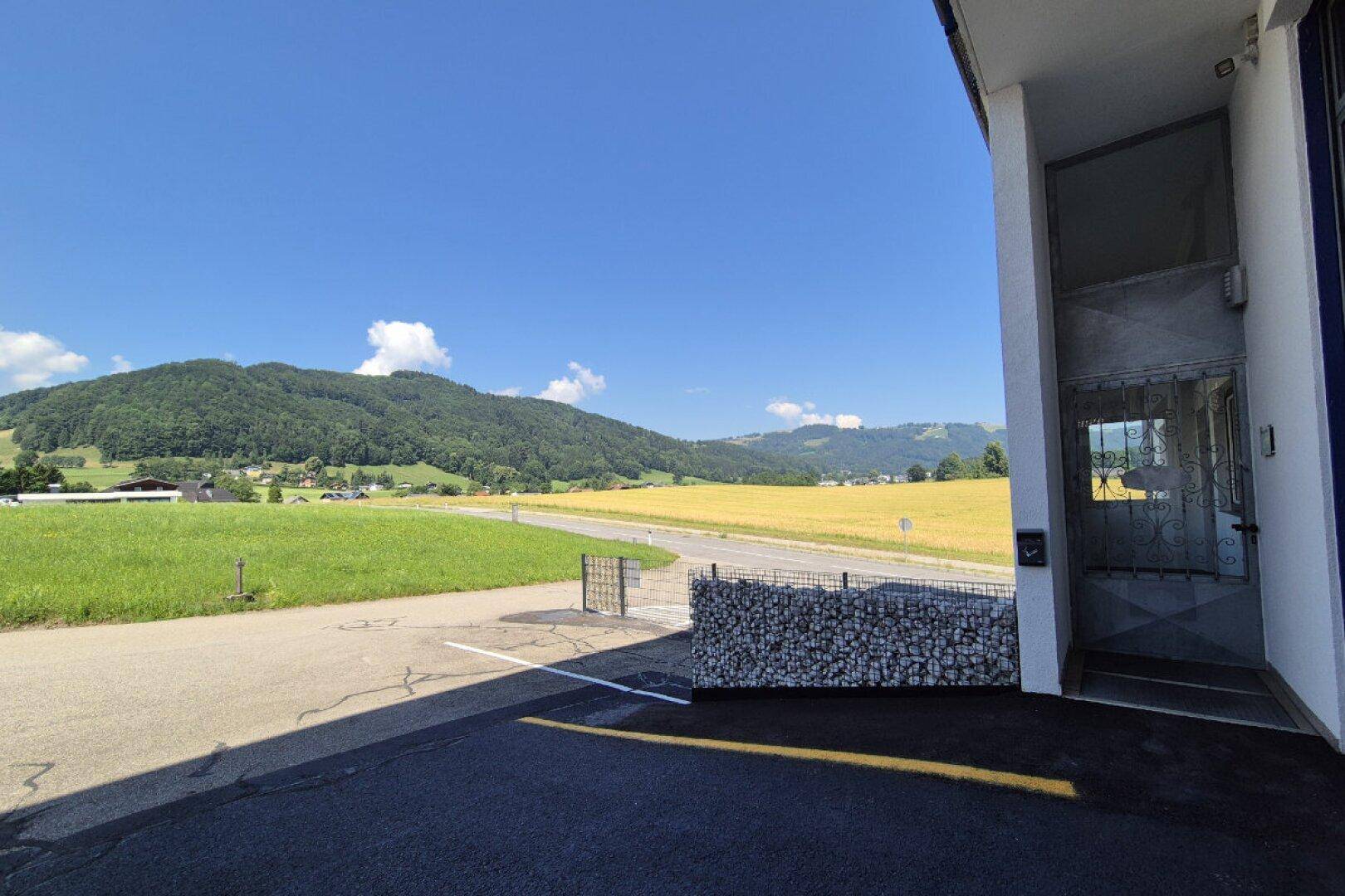 Außenansicht des Gebäudeeingangs mit Parkplatz, Gabionenwand und weitem Blick auf Felder und Hügel.