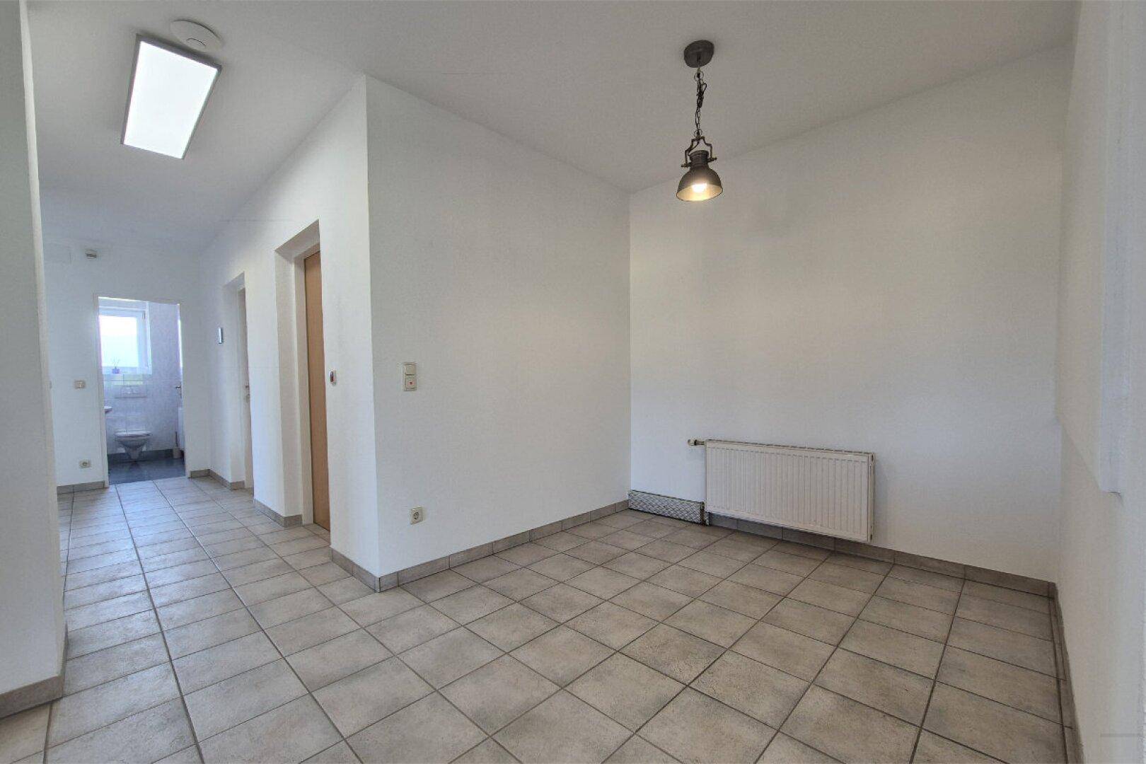 Empfangsbereich oder Wartezimmer mit Fliesenboden und einer einfachen Deckenleuchte, angrenzend an einen Flur.