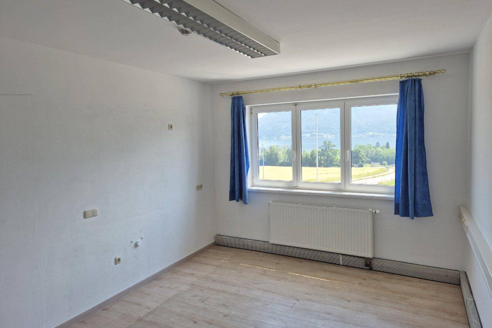 Leeres Büro mit hellem Laminatboden, Heizkörper und Fenster mit Seeblick und blauen Vorhängen.