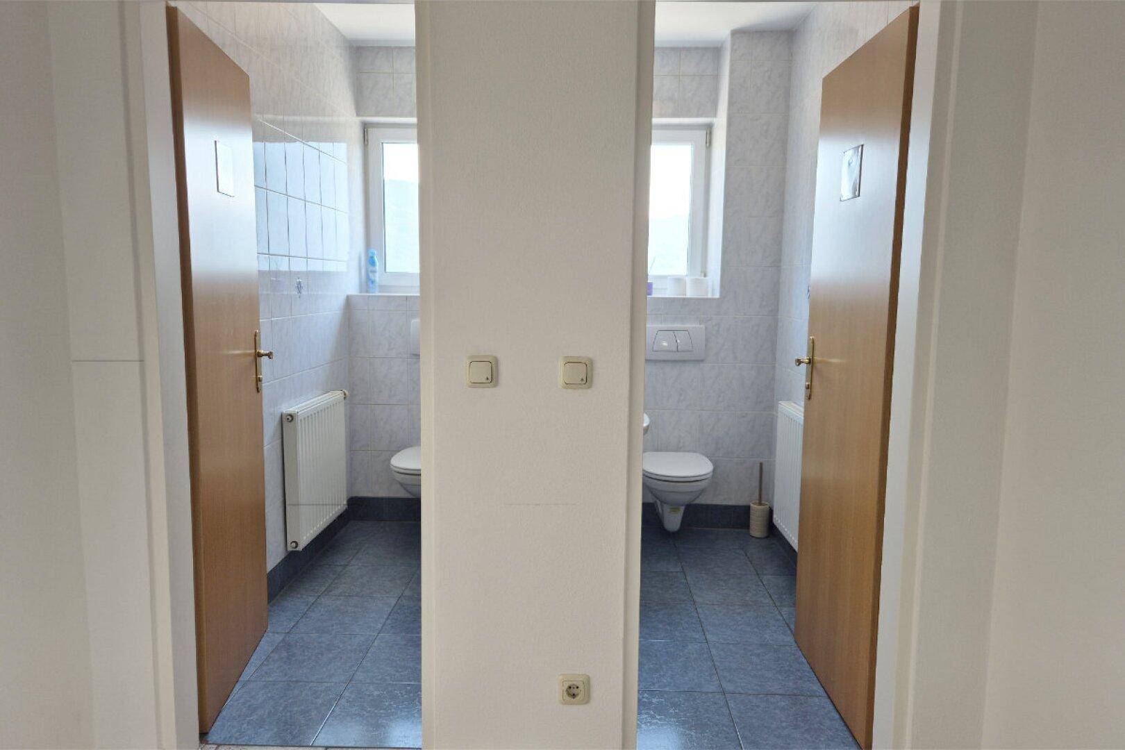 Zwei separate Toilettenräume mit weißen WCs, gefliesten Wänden und jeweils einem Fenster.