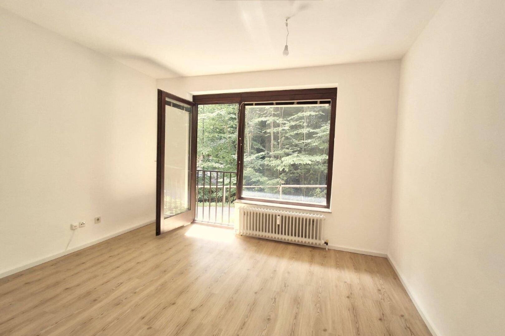Geräumiger Wohnbereich mit hellem Holzboden, großem Fenster und Zugang zum Balkon.