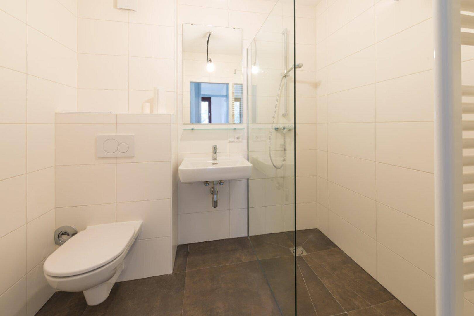 Modernes Badezimmer mit weißen Fliesen, wandhängendem WC und ebenerdiger Dusche mit Glaswand.