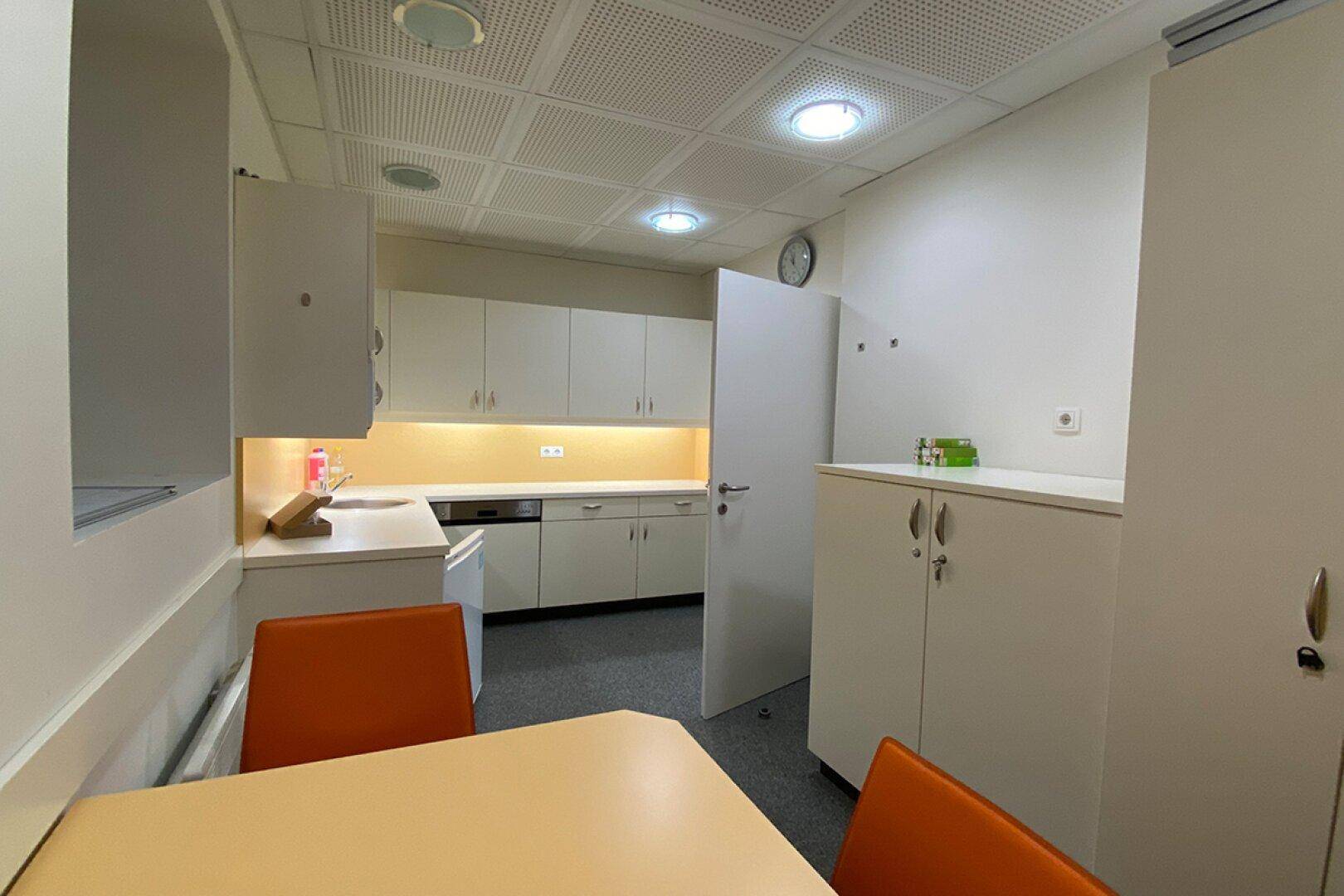 Kleine Küchenzeile oder Pausenraum mit weißen Schränken, Spüle und einem Tisch mit orangefarbenen Stühlen.