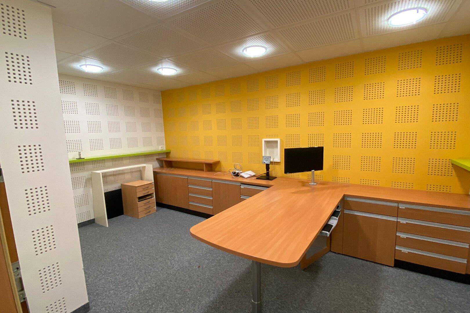 Büroraum mit einer gelben Akzentwand, Holzmöbeln und Teppichboden, gut beleuchtet durch Deckenleuchten.