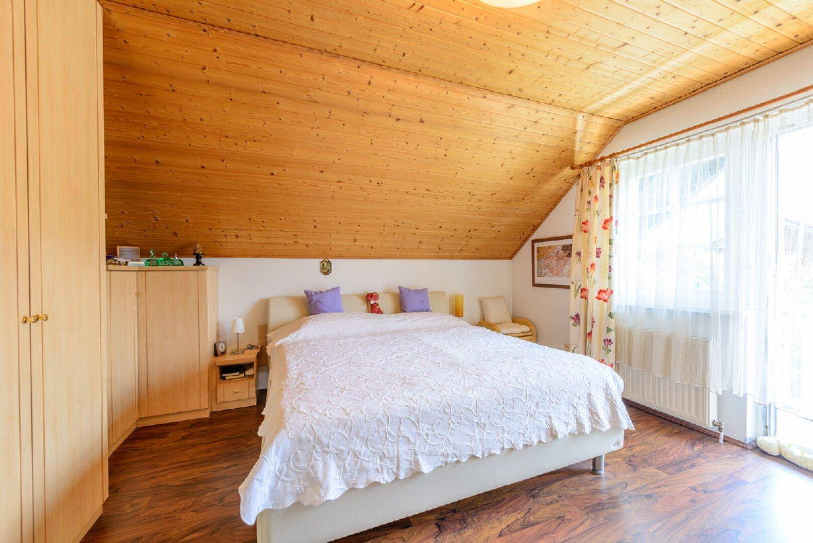 Schlafzimmer mit Holzbalkendecke, Holzboden und großem Fenster, das viel Licht hereinlässt.