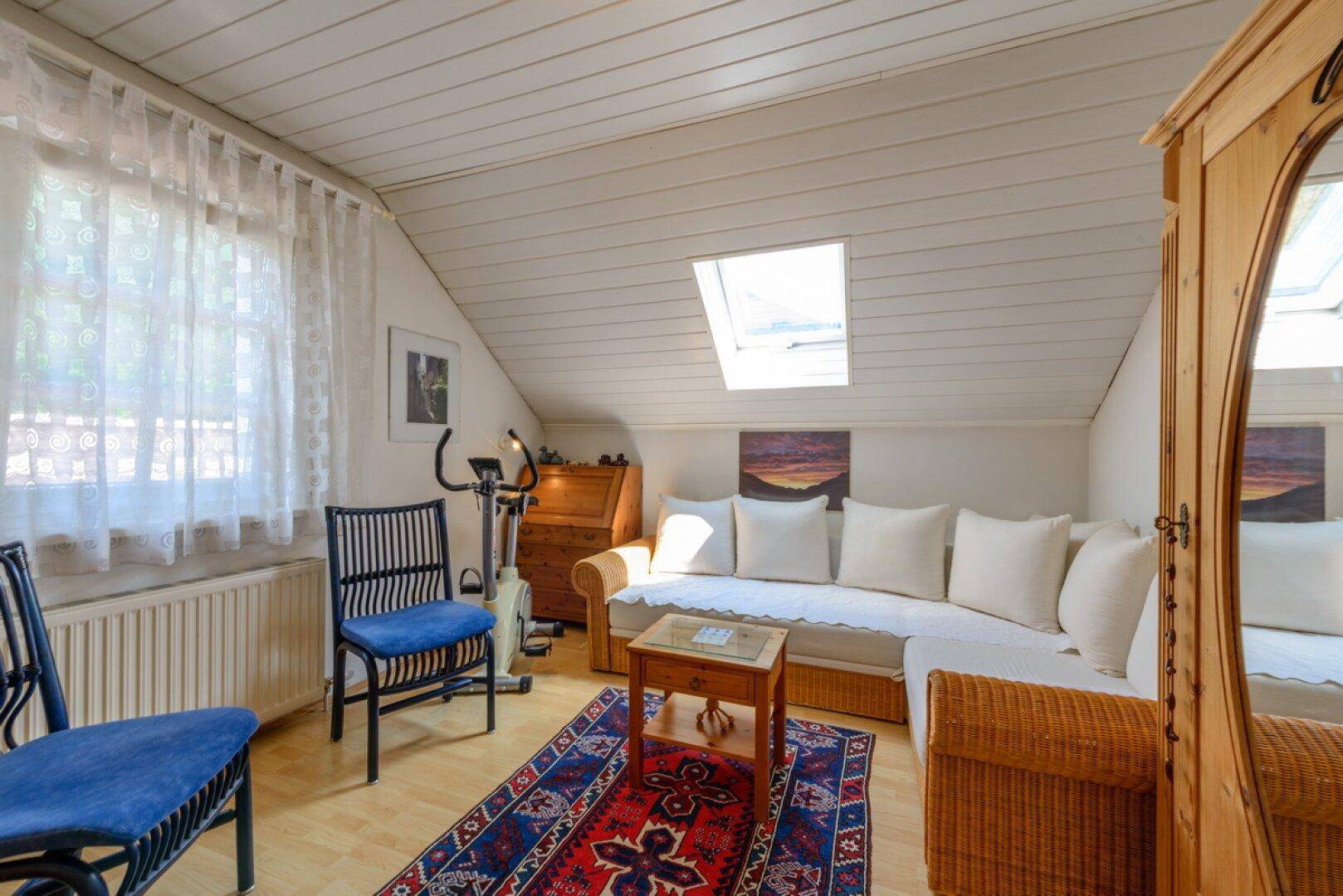 Vielseitiger Raum mit Sofa, Schreibtisch und Fitnessgerät unter einem Dachfenster.