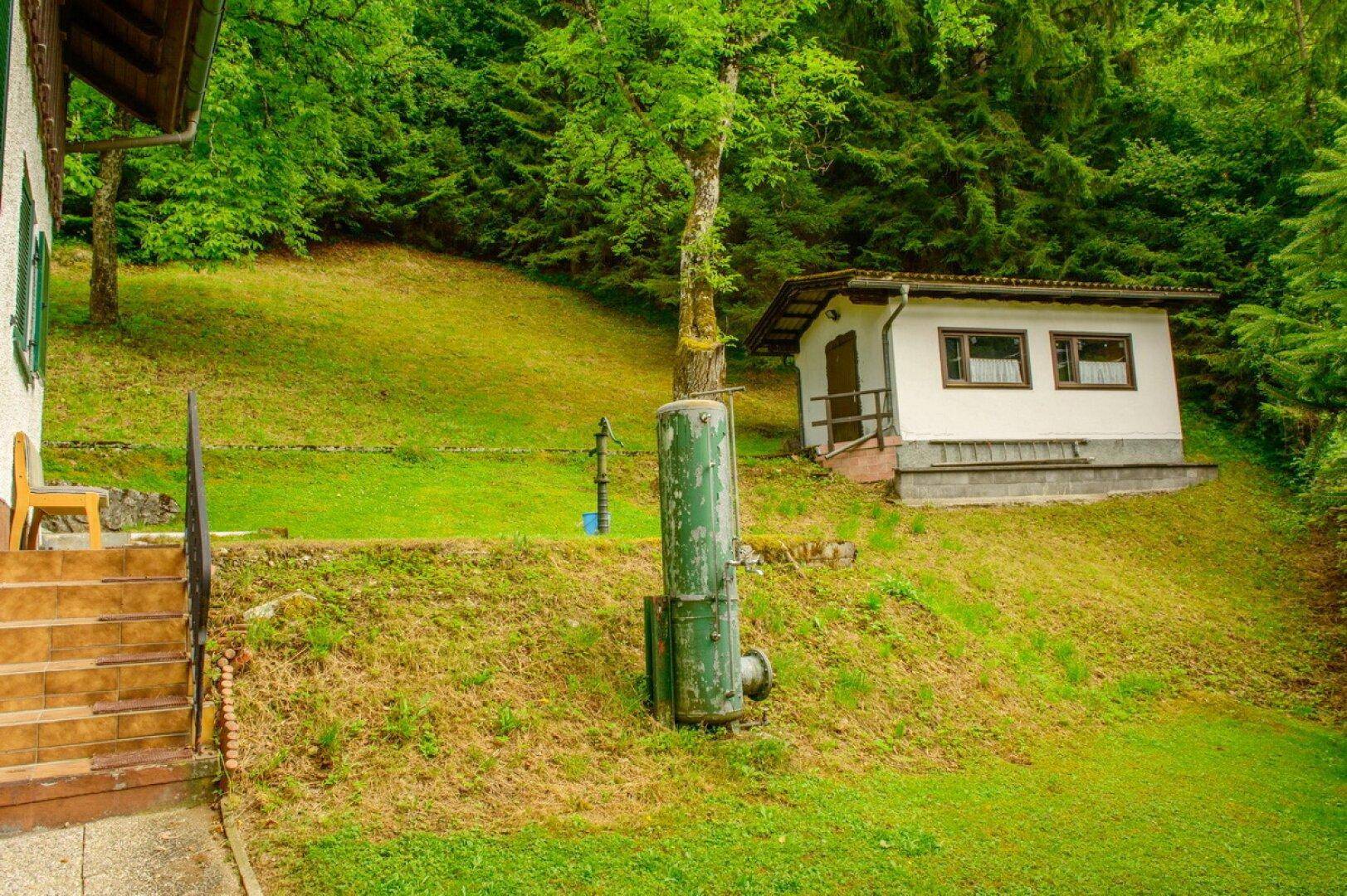 Kleines Nebengebäude auf einem grünen Hanggrundstück umgeben von Bäumen.