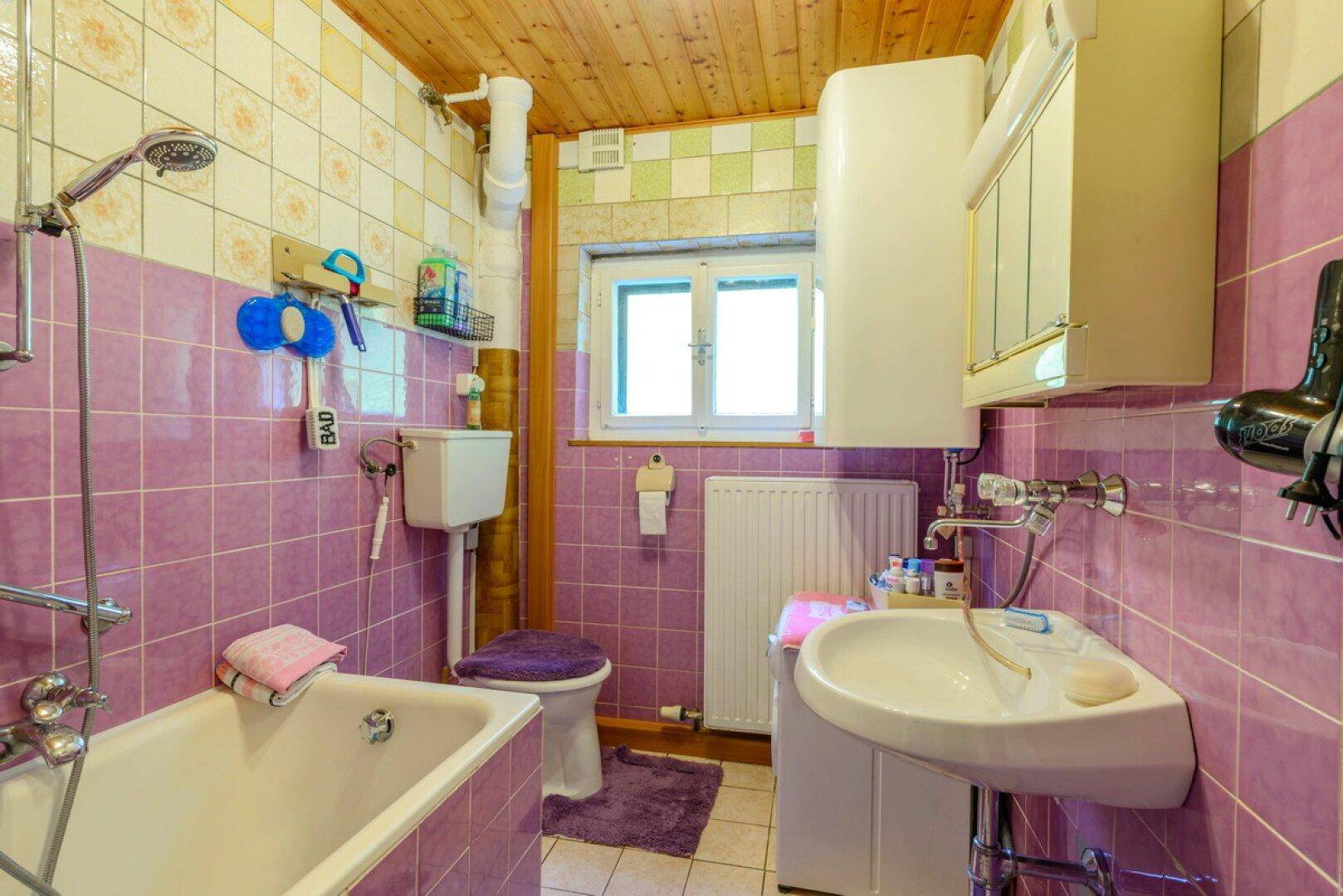 Badezimmer mit lila Fliesen, Badewanne, WC und Waschbecken, ausgestattet mit Holzdecke.
