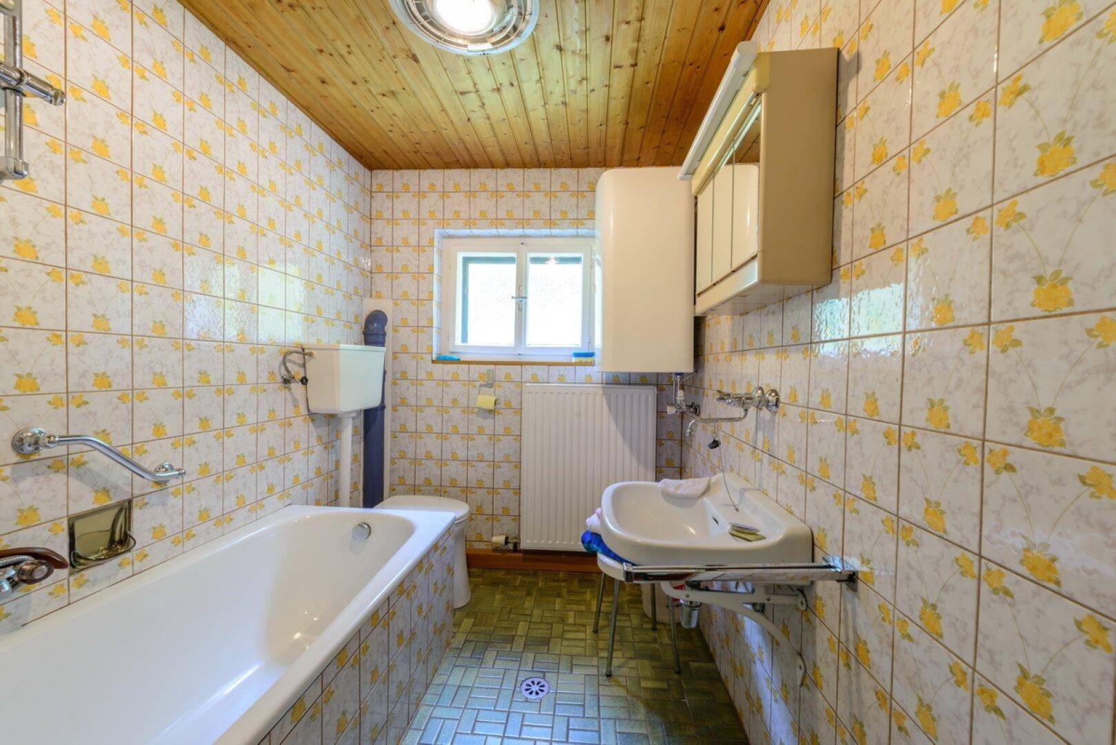 Badezimmer mit gelb gemusterten Fliesen, Badewanne, WC und Waschbecken, mit Holzdecke.