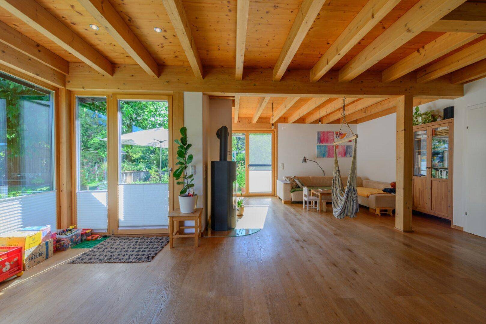 Heller Wohnbereich mit Holzbalkendecke, Holzboden, Kaminofen und großen Fensterfronten zum Garten.
