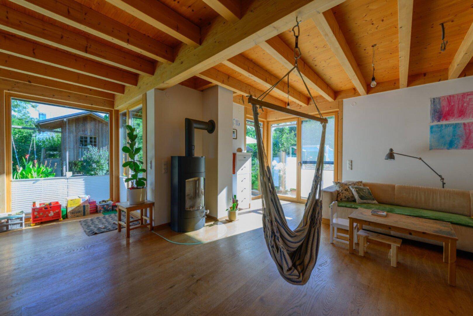 Gemütlicher Wohnbereich mit Kaminofen, Holzbalkendecke und Hängesessel, umgeben von großen Fenstern.