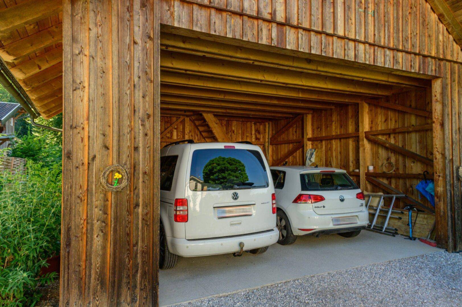 Überdachter Carport aus Holz bietet Platz für zwei Fahrzeuge, umgeben von grüner Vegetation.