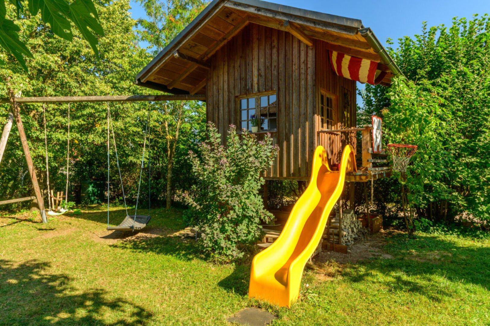 Spielbereich im Garten mit Baumhaus, gelber Rutsche und Schaukel für Kinder.