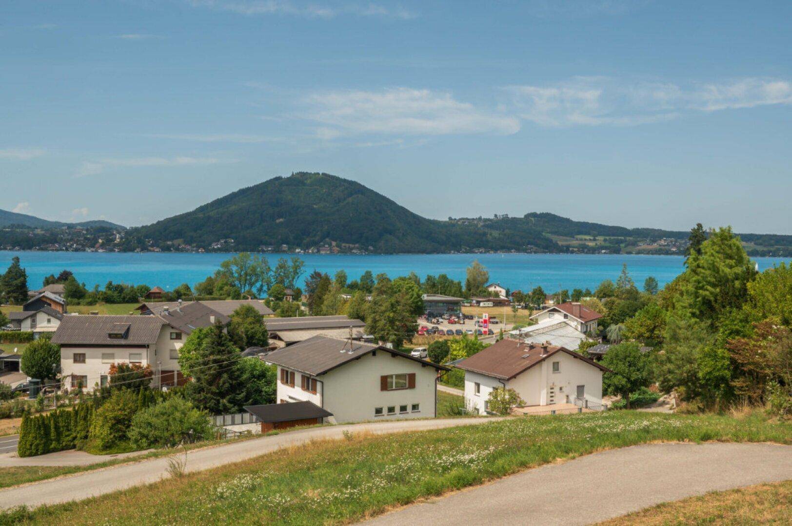 Blick auf eine Wohngegend mit Häusern, grünen Wiesen und dem Attersee im Hintergrund.