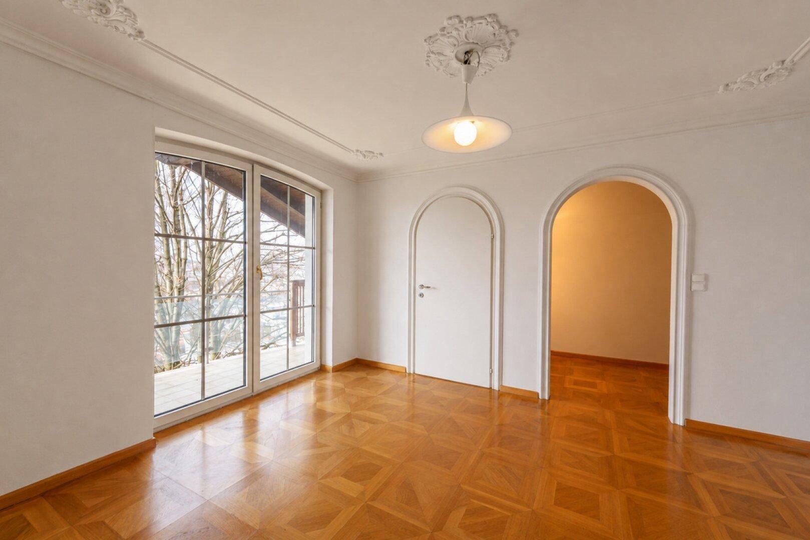 Heller Raum mit Fischgrätparkett, Stuckdecke und großem Fenster mit Zugang nach draußen.
