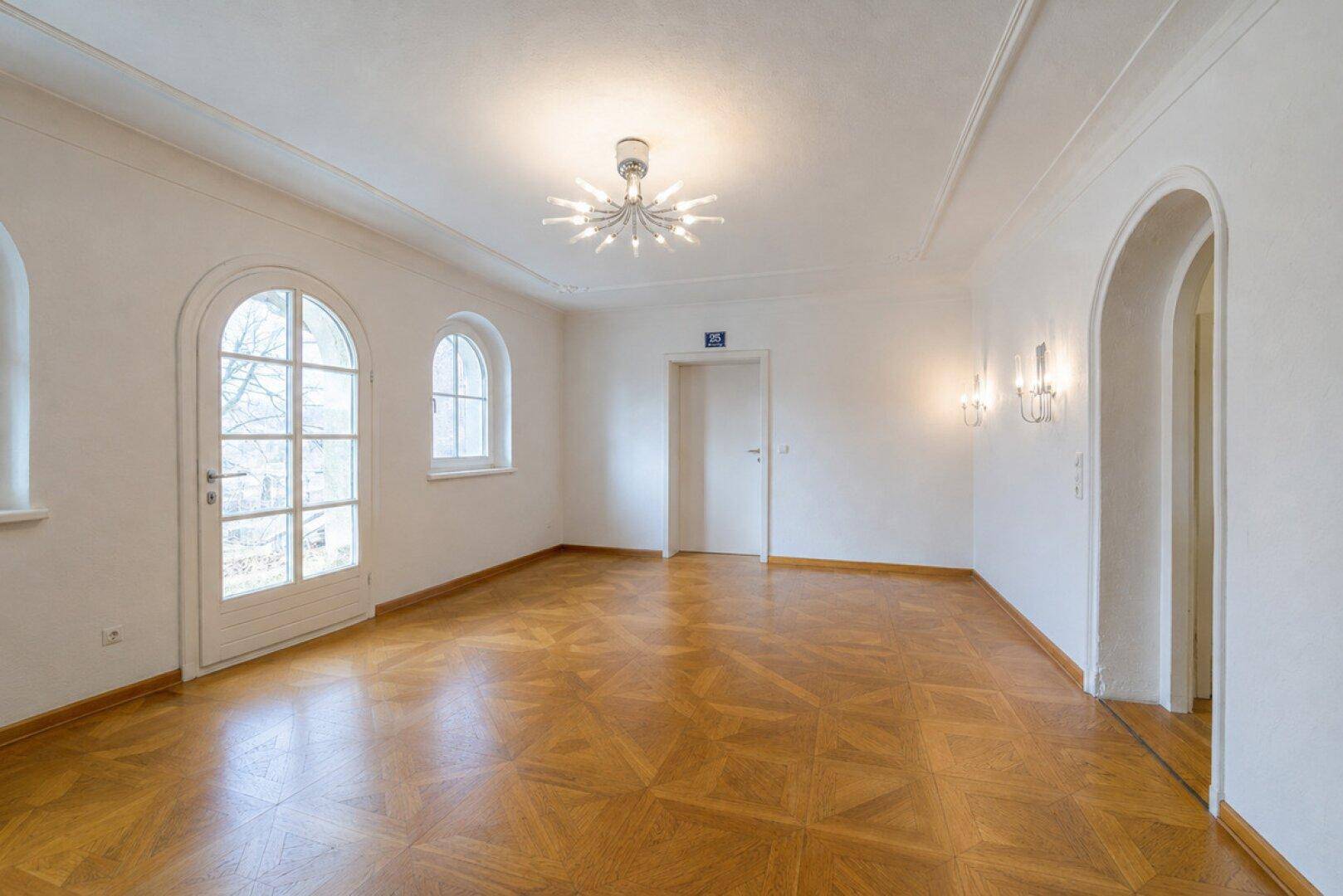 Geräumiger Wohnraum mit Parkettboden, Stuckdecke, modernen Leuchten und Bogenfenstern.