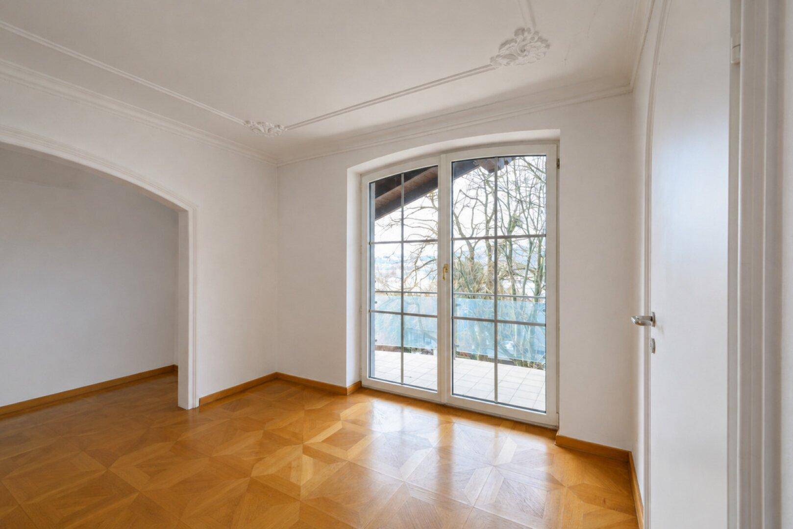 Heller Raum mit Parkettboden, Stuckdecke und großem Fenster mit Blick ins Grüne.