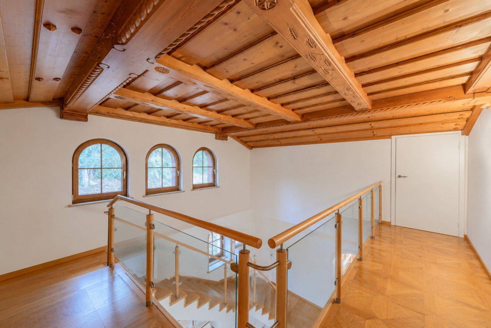 Obergeschoss mit Holztreppe, Glasgeländer, kunstvoller Holzdecke und Bogenfenstern.