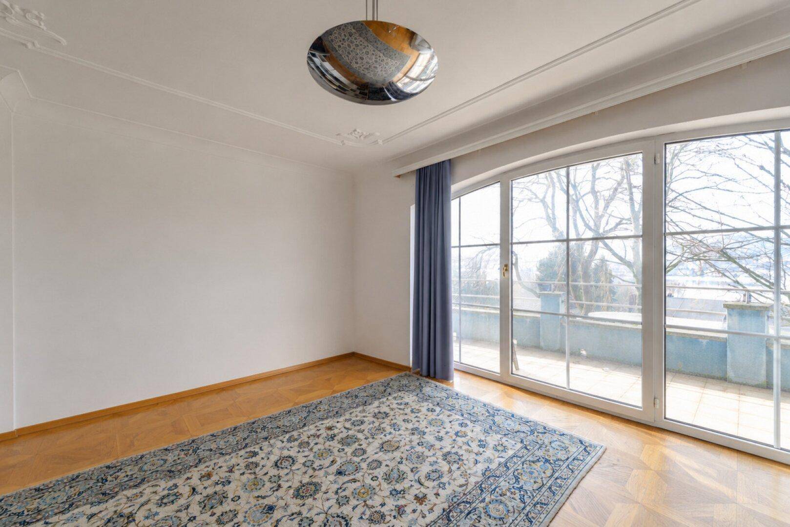Heller Raum mit Parkettboden, Stuckdecke und breiter Fensterfront mit Zugang zur Terrasse.