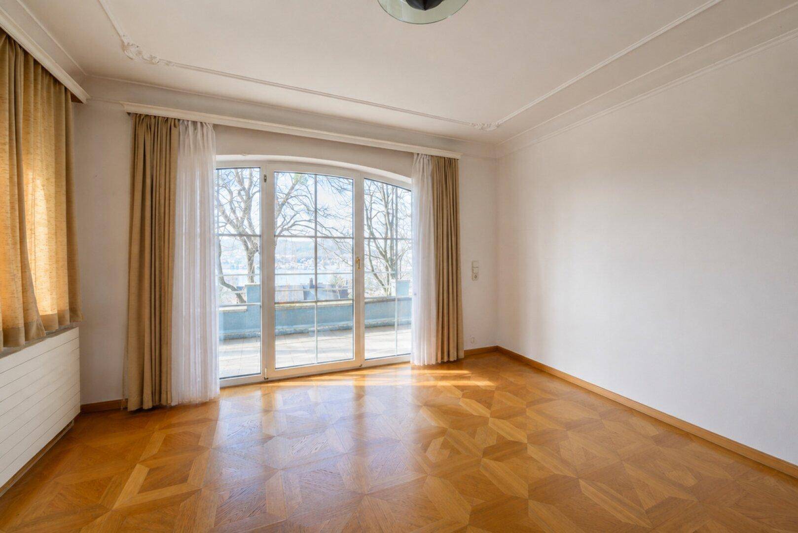 Geräumiger Raum mit Parkettboden, Stuckdecke und großer Fensterfront mit Zugang nach draußen.